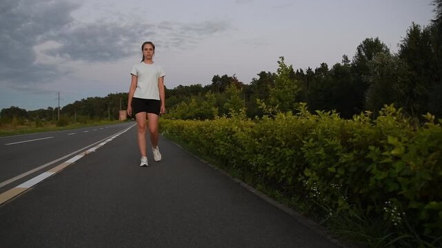 Young woman walking and then accelerating into running toward camera at sunset, filmed from low ground level in slow motion. Front tracking shot highlights transition, power, and dynamic urban fitness