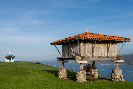 A stunning landscape featuring a traditional wooden h&oacute;rreo (granary) on stone pillars and the blue-and-white Regalina Chapel on a green clifftop overlooking the Cantabrian Sea in Cadavedo, Asturias, S