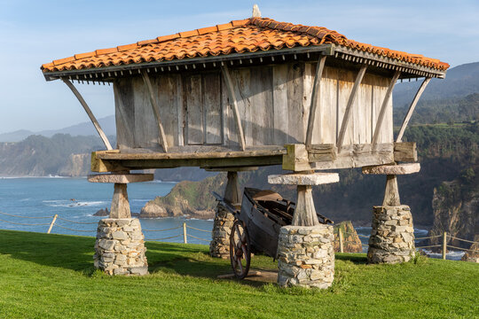 A stunning landscape featuring a traditional wooden h&oacute;rreo (granary) on stone pillars and the blue-and-white Regalina Chapel on a green clifftop overlooking the Cantabrian Sea in Cadavedo, Asturias, S