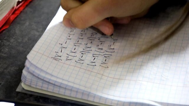 A student's hand diligently writes the process of solving a mathematical equation in a squared notebook using a blue pen. The footage demonstrates calculations, logical thinking, educational process, 