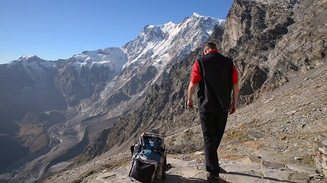 Beautiful summer day in the Italian Alps: hiker rests beside backpack at Sella Bivouac admiring Monte Rosa and its glaciers, with Alpe Pedriola and Lago delle Locce below, Macugnaga, Piedmont