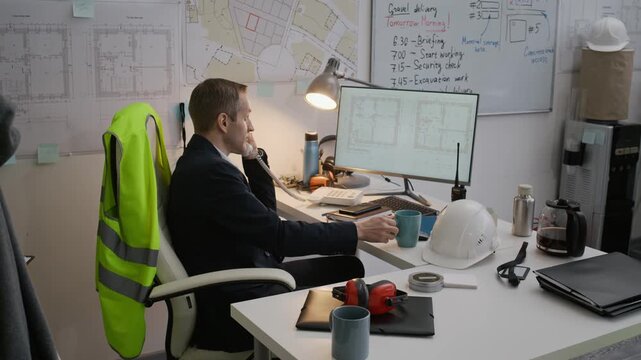 Arc shot of male Caucasian engineer in suit sitting at table while analyzing blueprint on computer and calling on landline phone in construction office