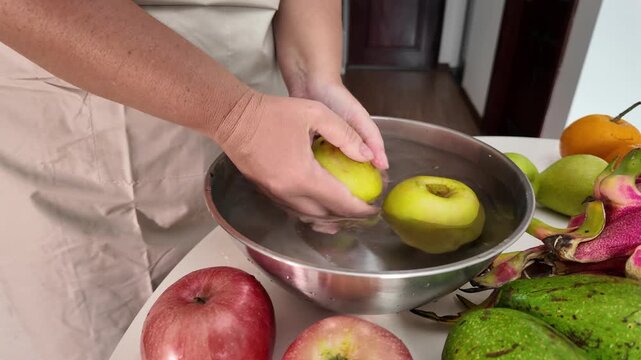 Woman's hands in an apron, standing in front of a table with fruit and washing green apples