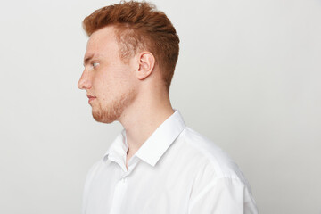 Profile of a young redhead man with beard wearing white shirt looking to the left on a plain light...
