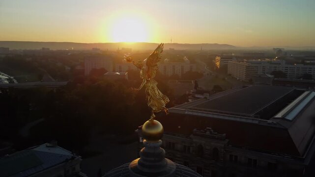 A circular aerial shoot of the statue of Pheme on the dome of the Academy of Arts in the Lipsiusbau building under sunshine at dawn with the Frauenkirche, Dresden's Old Town and the River Elbe behind 
