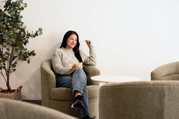 Fototapeta premium Relaxed young woman sitting in armchair in bright minimalist living room, soft daylight and calm atmosphere representing comfort, balance and modern lifestyle.