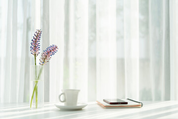 White coffee cup and a vase of Bromeliad Matchstick plant, phone and notebook, on white table