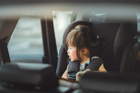 Happy little Asian girl sitting safely in car seat and looking out the window, toddler secured in child safety seat for road trip, concept of family travel, passenger safety.