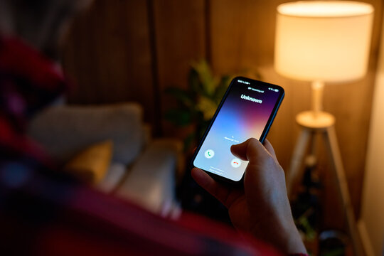 Close-up of hand holding smartphone showing unknown incoming call in living room. Concept of spam call, telemarketing, scam risk and mobile communication