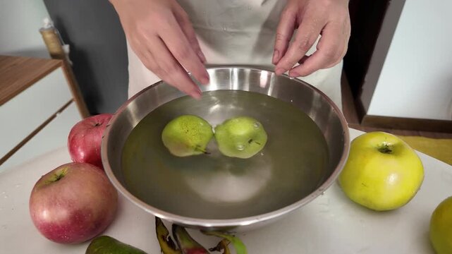 The hands of a woman in an apron, standing in front of a table with fruit and washing ripe pears in a metal bowl of water