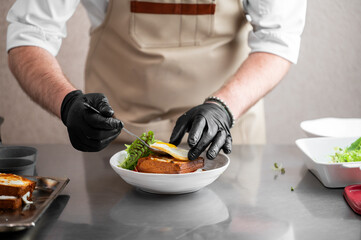 A professional chef in a tan apron and black nitrile gloves uses tweezers to carefully plate a...
