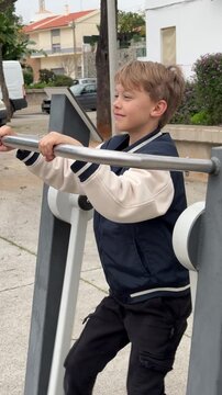 Smiling school aged boy in a varsity jacket enjoying a workout on an elliptical machine at an outdoor public gym, promoting a healthy and active lifestyle for children in a city park setting