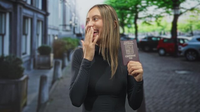 Young woman holding czech passport and covering mouth with hand while smiling on a cobbled street; travel excitement.