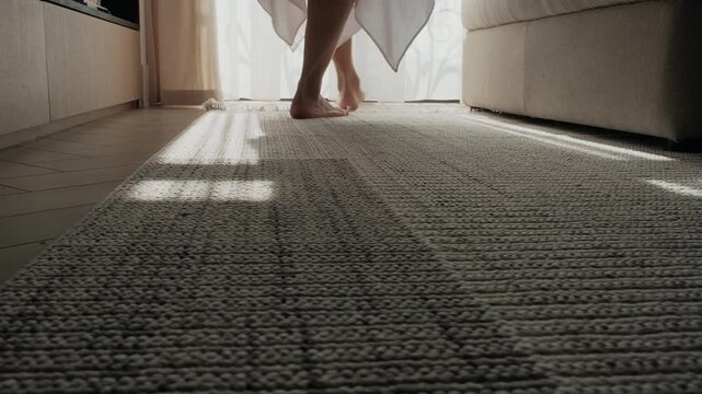 Barefoot woman walking across a rug in a modern apartment. Close-up of feet, calm home lifestyle scene in a minimalist residential interior. Neutral color palette, modern furniture in a soft morning