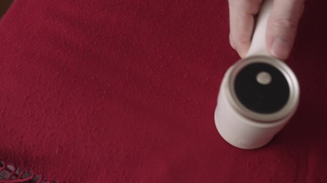 Top view of male hand operating electric lint remover on red wool scarf, effectively cleaning fabric by shedding pilling and restoring smooth surface, symbolizing garment care, close-up, slow motion.