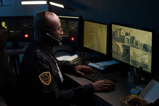 Black man wearing headset working as security officer monitoring multiple surveillance screens in control room at night, focusing on urban building footage and maps