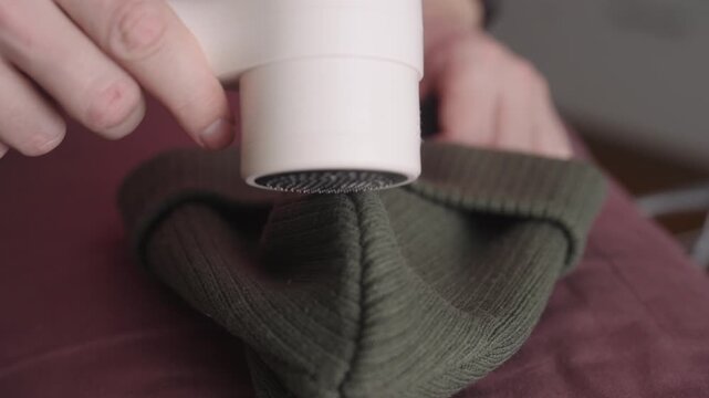 Man using electric lint remover machine on green knitted winter hat, refreshing textile fabric care and improving worn clothing appearance. Concept of effective fabric care and maintenance, closeup.
