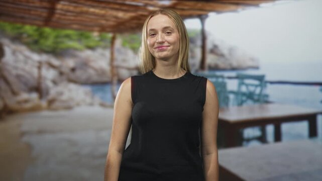 Woman with bare arm, eyes closed and smiling while standing by a seaside cafe table under wooden pergola near rocky cove; serene leisure.
