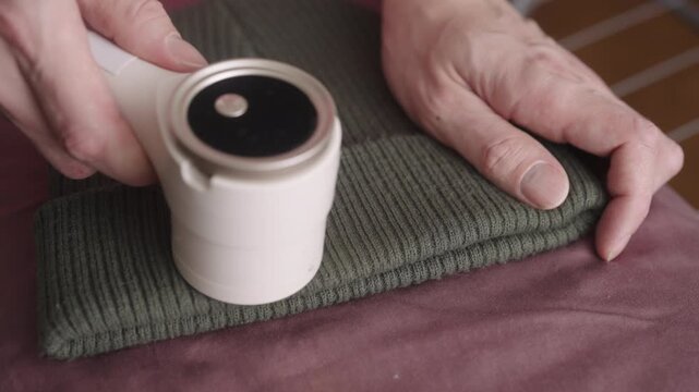 Hand applying electric lint remover to green knitted beanie, efficiently defuzzing textile material and restoring fabric surface by removing pills and bobbles for renewed garment quality, close-up.