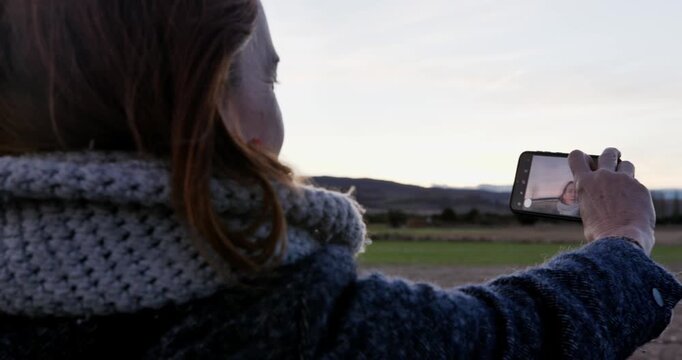 Capturing the Moment: A woman, captured from behind, immerses herself in a moment, using a smart device to immortalize a picturesque landscape.
