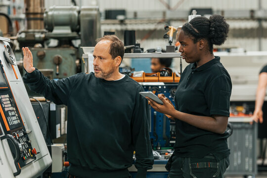 Female worker taking notes while coworker explaining machine parts in factory