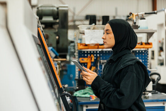 Young female worker wearing hijab operating machine with digital tablet in factory