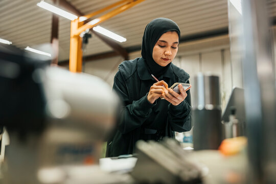Low angle view of female factory worker wearing hijab taking notes on note pad