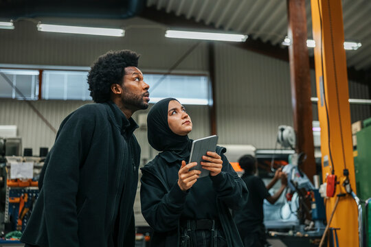 Male worker standing near female coworker operating machine with digital tablet in factory
