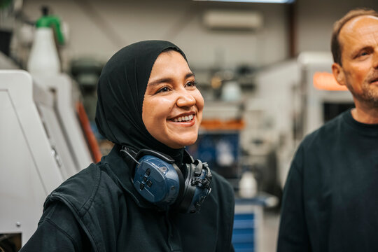 Smiling female worker wearing hijab with ear protectors around neck in factory