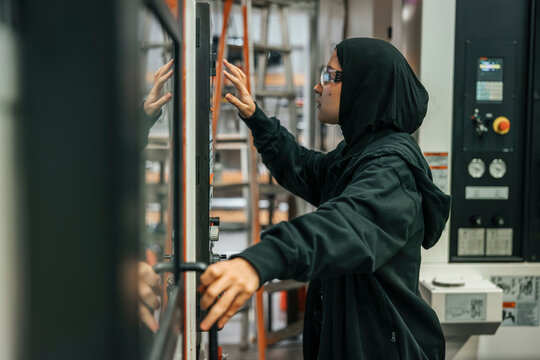 Side view of focused young female worker wearing hijab operating machine in factory