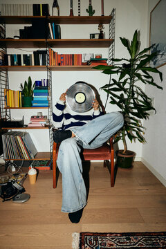 Young man covering face with vinyl record while sitting on chair at home