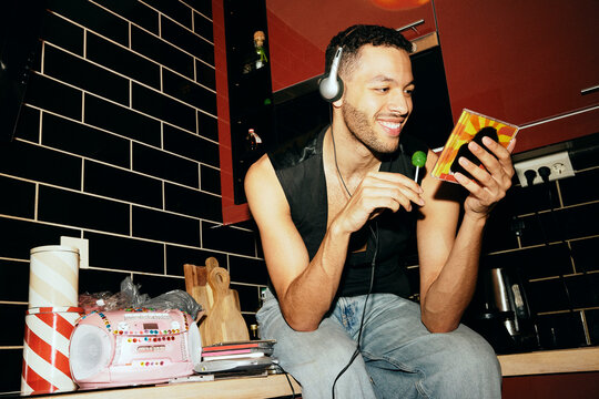 Low angle view of smiling man holding reading cover art and listening to music while sitting on kitchen counter at home