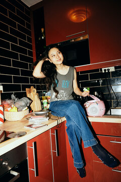 Smiling young woman with hand in hair sitting on kitchen counter at home