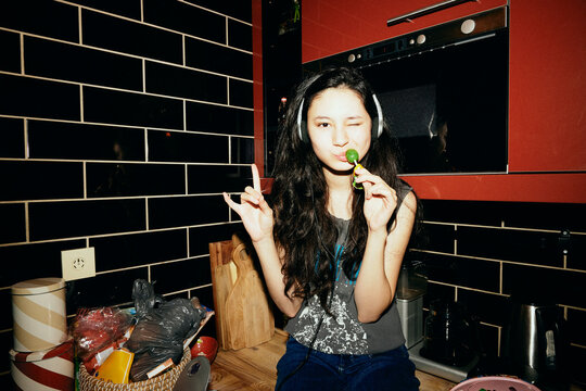 Portrait of young woman making horn sign while listening to music through headphones in kitchen at home