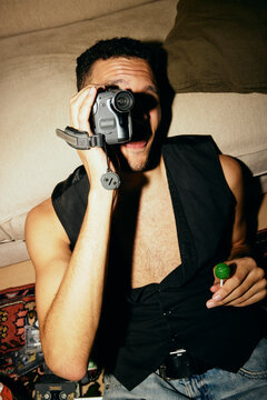 Young man holding candy while looking through vintage video camera at home