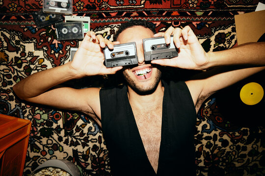 Directly above shot of young man covering eyes with audio cassettes while lying on carpet at home