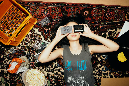 Directly above shot of woman covering eyes with audio cassettes while lying on carpet at home