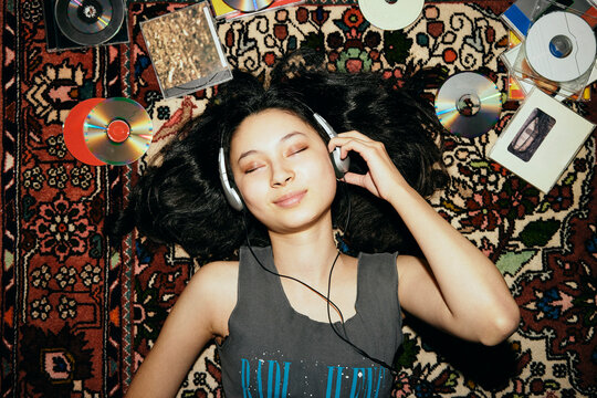 Directly above shot of young woman listening to music with eyes closed while lying amidst CDs on carpet at home