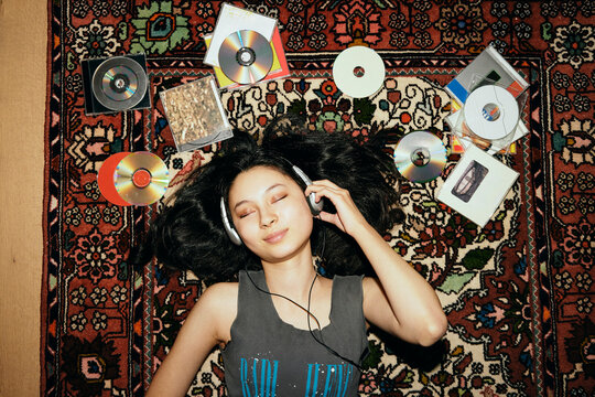 Smiling young woman listening to music through headphones while lying on carpet amidst CDs