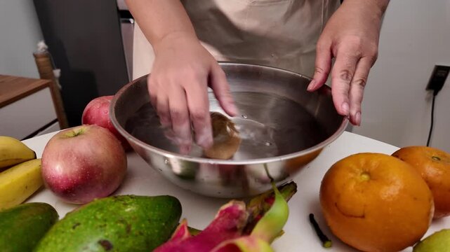 Close-up of a woman's hands in an apron, standing in front of a table with fruit and washing a kiwi