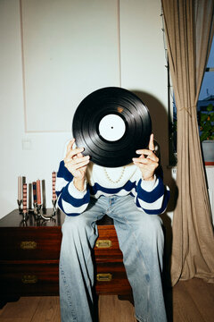 Man covering face with record vinyl while sitting on table at home