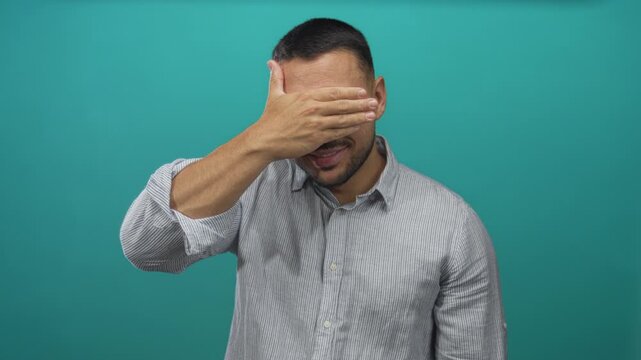 Young hispanic man covers eyes with hand and smiles broadly against a bright teal studio wall; playful shyness.