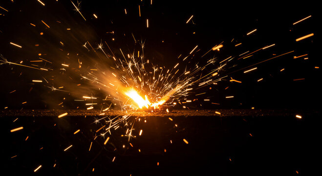 Orange sparks bursting from metal grinding. Intense fire effects in dark environment. Industrial welding and fabrication process visualization for manufacturing and engineering concepts.