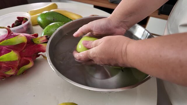 The hands of a woman in an apron standing in front of a table with fruit and washing green apples