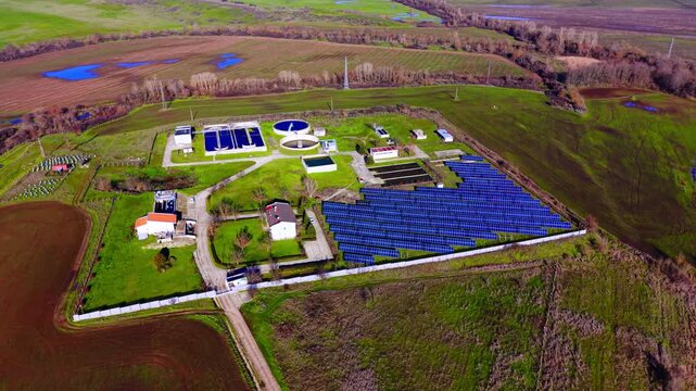 Water purification facility and photovoltaic station in a rural green field under clear sky. Aerial view of wastewater treatment plant with solar panel farm.