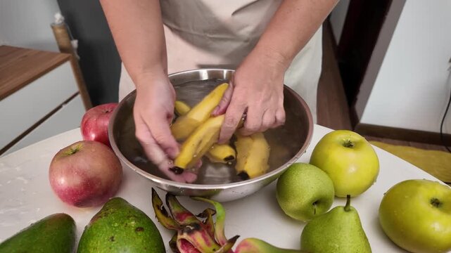 The hands of a woman in an apron standing in front of a table with fruit and washing bananas