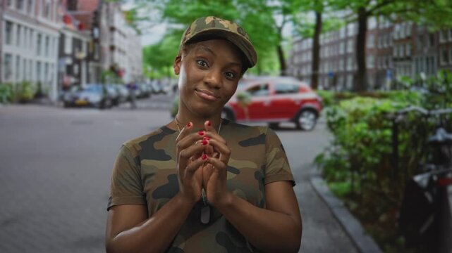 Woman in camouflage uniform clasps dogtags and folds hands on a street with parked cars and brick buildings; readiness determination.
