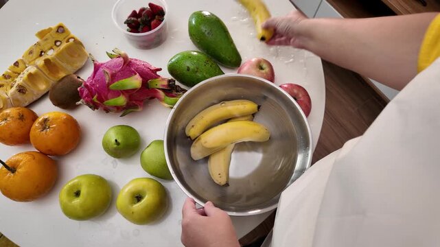 Hands of a woman in an apron standing in front of a table with fruit and washing bananas