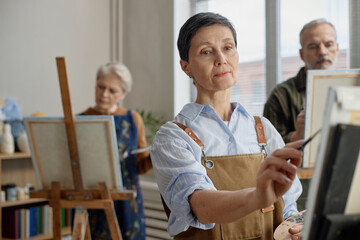 Obraz premium Middle aged Caucasian woman painting on canvas in classroom setting, standing in foreground with focused expression, senior Caucasian woman and senior Caucasian man painting in background
