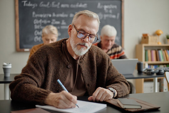 Middle aged Caucasian man wearing glasses sitting at desk, writing in notebook during classroom lesson with senior Caucasian woman and senior Caucasian man studying in background